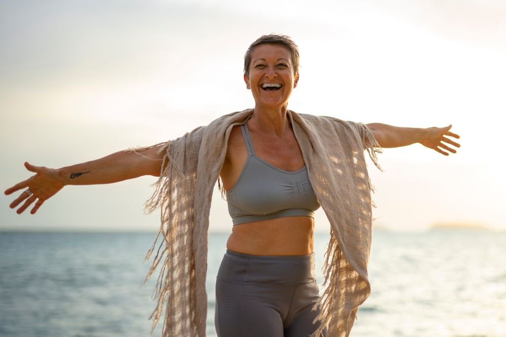 Menopausa: Como evitar a perca de massa óssea, controlar o peso e ganhar vitalidade com Exercício Físico -mulher de braços abertos e sorrindo na praia