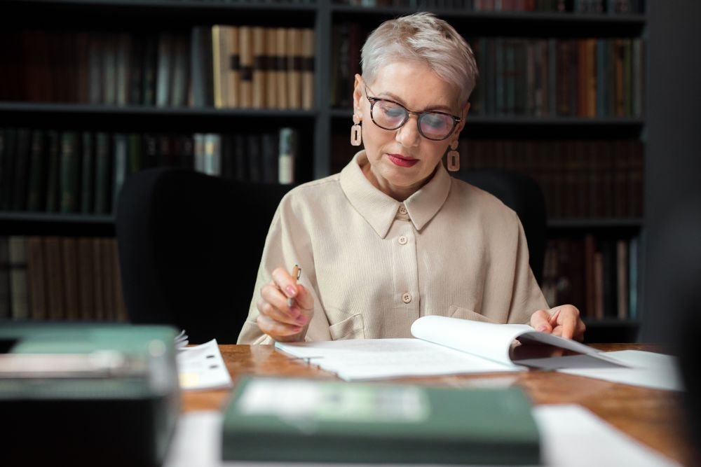 mulher madura lendo em uma mesa com vários livros, onde compara as histórias. Isso a ajuda na construção do conhecimento.